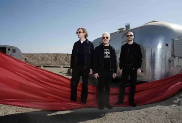 Le trio Above & Beyond pose devant deux caravanes argentées dans un paysage désertique, vêtu de noir et entouré d'un long voile rouge qui traverse le premier plan de la scène.