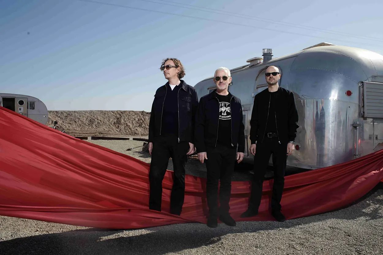 Le trio Above & Beyond pose devant deux caravanes argentées dans un paysage désertique, vêtu de noir et entouré d'un long voile rouge qui traverse le premier plan de la scène.