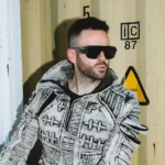 Gareth Emery avec une veste texturée grise et des lunettes de soleil noires devant un conteneur industriel, dans une ambiance urbaine et contemporaine.