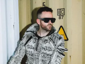 Gareth Emery avec une veste texturée grise et des lunettes de soleil noires devant un conteneur industriel, dans une ambiance urbaine et contemporaine.