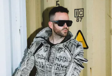 Gareth Emery avec une veste texturée grise et des lunettes de soleil noires devant un conteneur industriel, dans une ambiance urbaine et contemporaine.