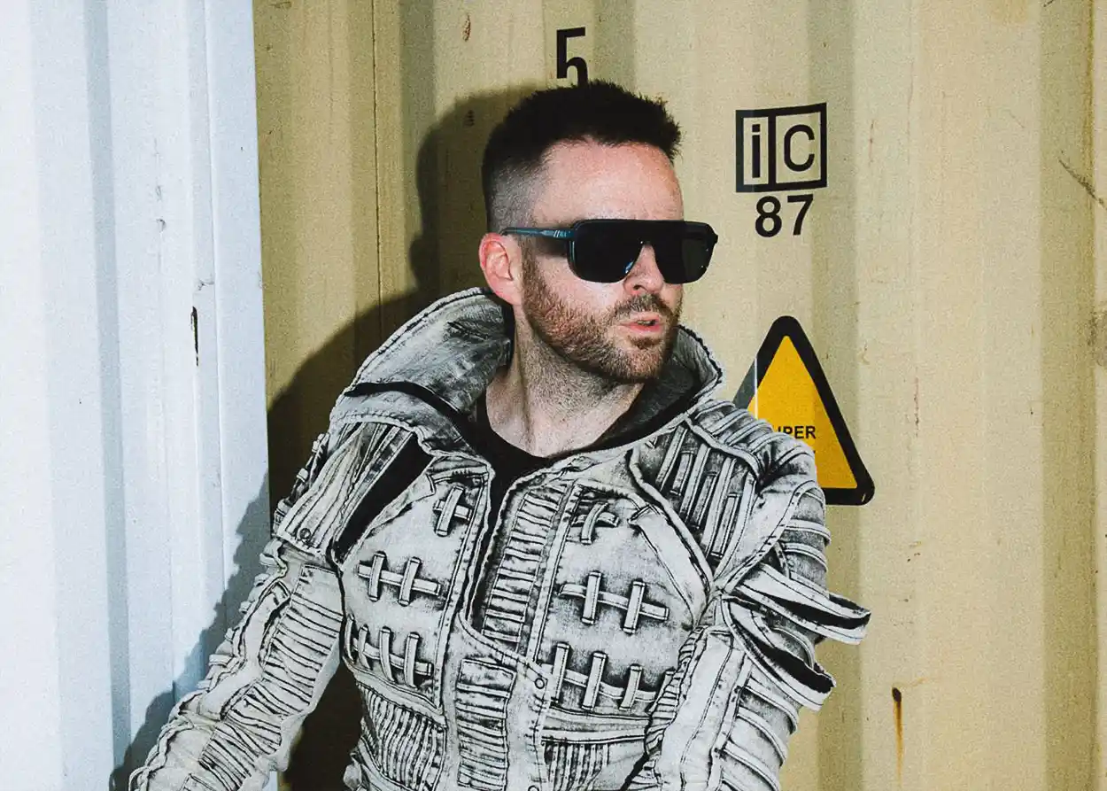 Gareth Emery avec une veste texturée grise et des lunettes de soleil noires devant un conteneur industriel, dans une ambiance urbaine et contemporaine.