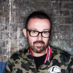 Judge Jules portant des lunettes noires et une veste camouflage, photographié devant un mur de briques au style industriel.