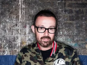 Judge Jules portant des lunettes noires et une veste camouflage, photographié devant un mur de briques au style industriel.
