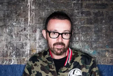 Judge Jules portant des lunettes noires et une veste camouflage, photographié devant un mur de briques au style industriel.