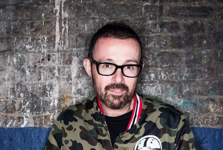 Judge Jules portant des lunettes noires et une veste camouflage, photographié devant un mur de briques au style industriel.