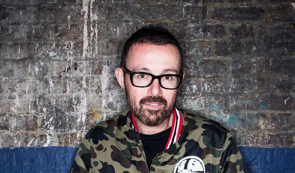 Judge Jules portant des lunettes noires et une veste camouflage, photographié devant un mur de briques au style industriel.