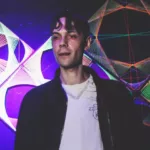 Stanley Fox se tenant derrière les platines dans une salle de concert, devant des décorations lumineuses aux couleurs violettes et multicolores, vêtu d'une veste noire sur un haut blanc.