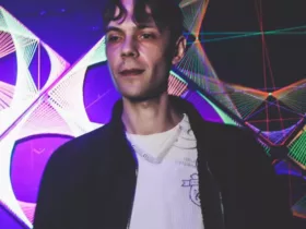 Stanley Fox se tenant derrière les platines dans une salle de concert, devant des décorations lumineuses aux couleurs violettes et multicolores, vêtu d'une veste noire sur un haut blanc.