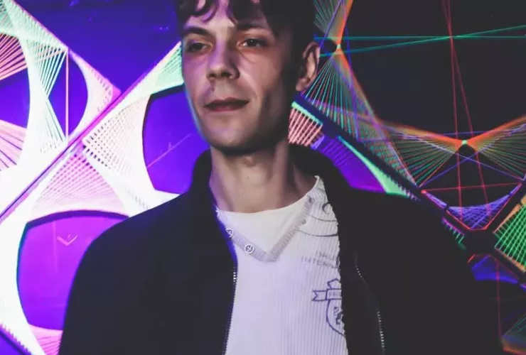 Stanley Fox se tenant derrière les platines dans une salle de concert, devant des décorations lumineuses aux couleurs violettes et multicolores, vêtu d'une veste noire sur un haut blanc.