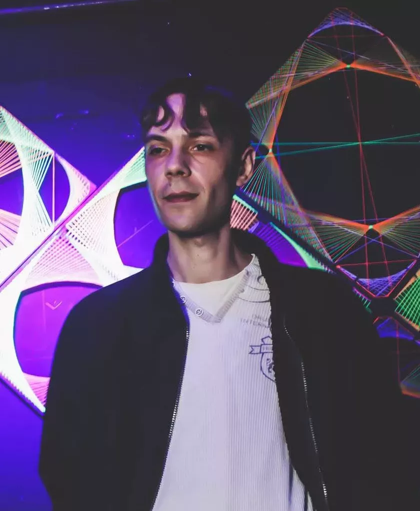 Stanley Fox se tenant derrière les platines dans une salle de concert, devant des décorations lumineuses aux couleurs violettes et multicolores, vêtu d'une veste noire sur un haut blanc.