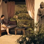 Paul van Dyk est assis avec un ordinateur portable et un casque audio dans un espace de détente du Château Bonheur Musique, avec vue sur la nature et une statue de Bouddha à proximité.