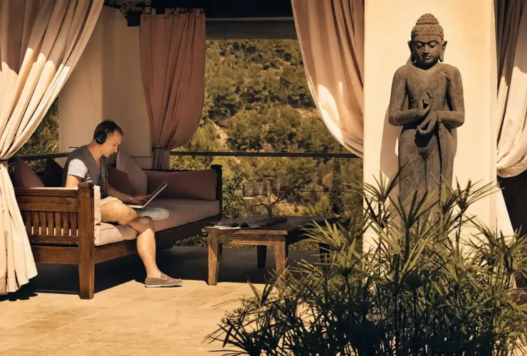 Paul van Dyk est assis avec un ordinateur portable et un casque audio dans un espace de détente du Château Bonheur Musique, avec vue sur la nature et une statue de Bouddha à proximité.