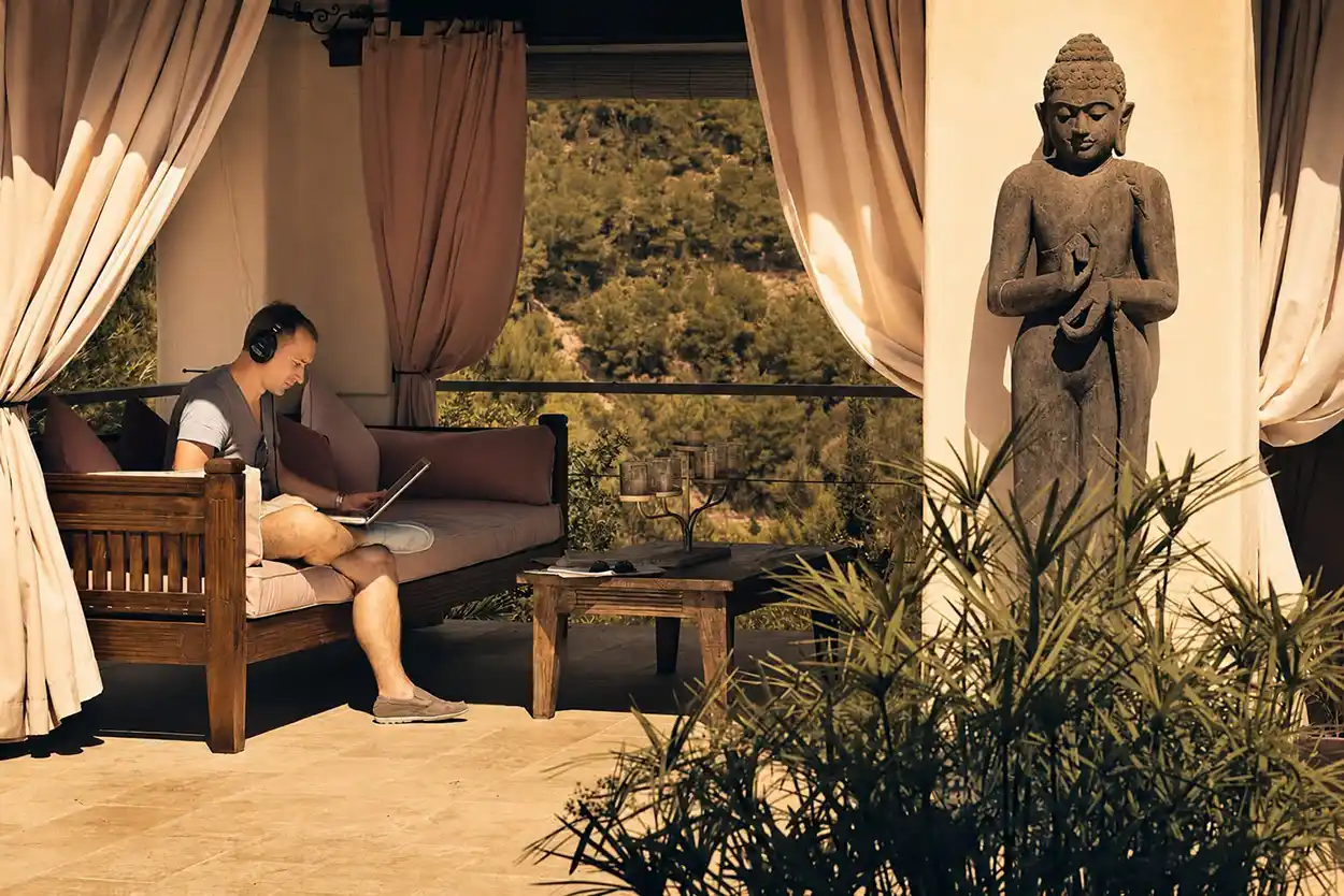 Paul van Dyk est assis avec un ordinateur portable et un casque audio dans un espace de détente du Château Bonheur Musique, avec vue sur la nature et une statue de Bouddha à proximité.