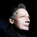 Portrait de William Orbit sur fond noir, le visage éclairé par une lumière vive tandis qu’il regarde vers le haut.