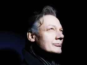 Portrait de William Orbit sur fond noir, le visage éclairé par une lumière vive tandis qu’il regarde vers le haut.