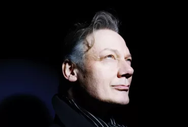 Portrait de William Orbit sur fond noir, le visage éclairé par une lumière vive tandis qu’il regarde vers le haut.