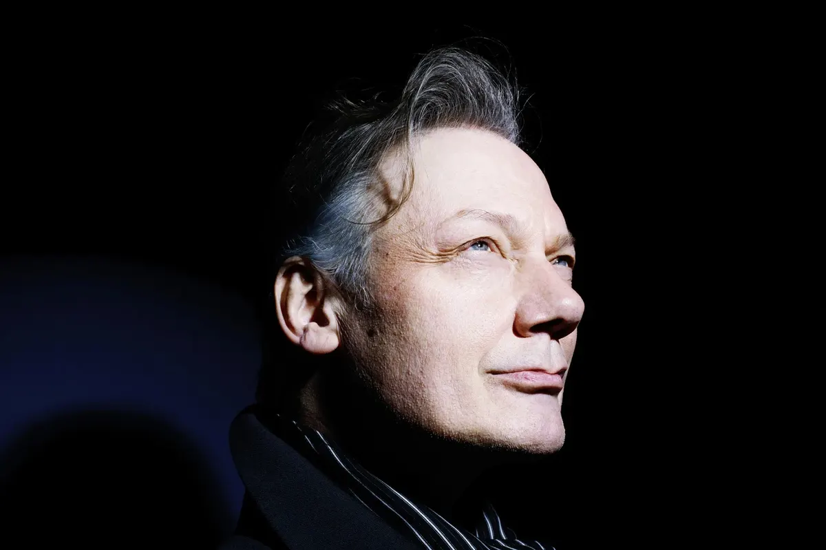 Portrait de William Orbit sur fond noir, le visage éclairé par une lumière vive tandis qu’il regarde vers le haut.