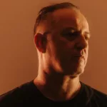 Portrait de Sasha dans une lumière chaude orangée, vêtu de noir et regardant légèrement sur le côté.