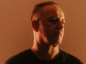 Portrait de Sasha dans une lumière chaude orangée, vêtu de noir et regardant légèrement sur le côté.