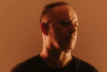 Portrait de Sasha dans une lumière chaude orangée, vêtu de noir et regardant légèrement sur le côté.