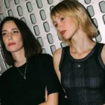 Amelie Lens et Angèle posent côte à côte, vêtues de noir, devant un décor métallique.