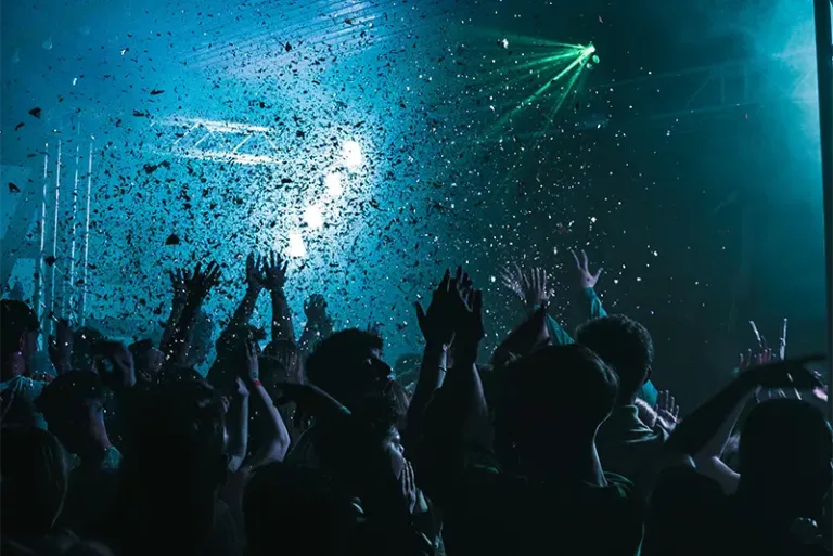 Foule  lors  d’une  soirée  de  musique  électronique,  les  bras  levés  sous  une  pluie  de  confettis  et  des  lumières  bleues  et  vertes.