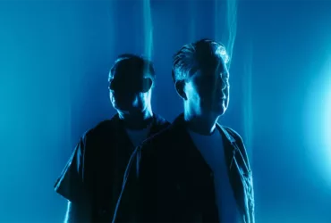 Sasha et John Digweed posent dans une lumière bleue intense, photographiés de trois quarts dans une ambiance sombre et atmosphérique pour “Northern Exposure Redux”.