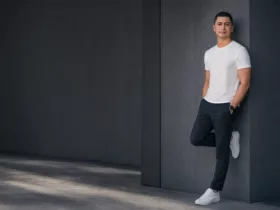 Portrait de Sylvermay en 2026, vêtu d’un t-shirt blanc et d’un pantalon sombre, adossé à un mur gris dans un décor architectural minimaliste.