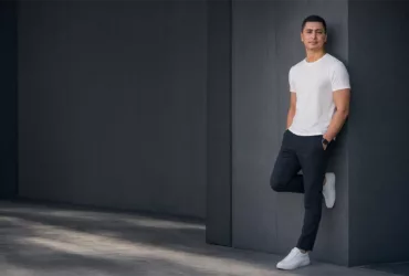 Portrait de Sylvermay en 2026, vêtu d’un t-shirt blanc et d’un pantalon sombre, adossé à un mur gris dans un décor architectural minimaliste.