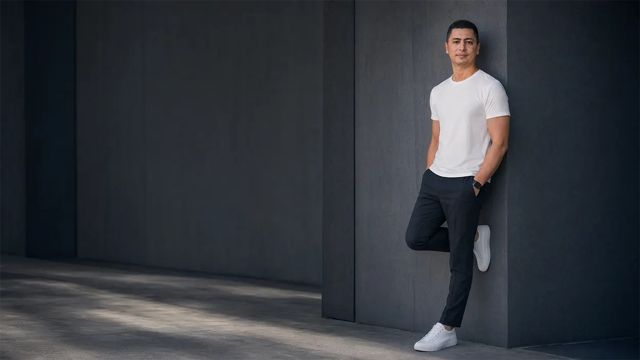 Portrait de Sylvermay en 2026, vêtu d’un t-shirt blanc et d’un pantalon sombre, adossé à un mur gris dans un décor architectural minimaliste.
