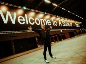 Armin van Buuren pose les bras ouverts devant une immense bannière « Welcome to A State Of Trance », dans une salle encore vide.