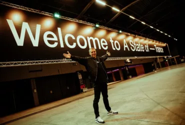 Armin van Buuren pose les bras ouverts devant une immense bannière « Welcome to A State Of Trance », dans une salle encore vide.