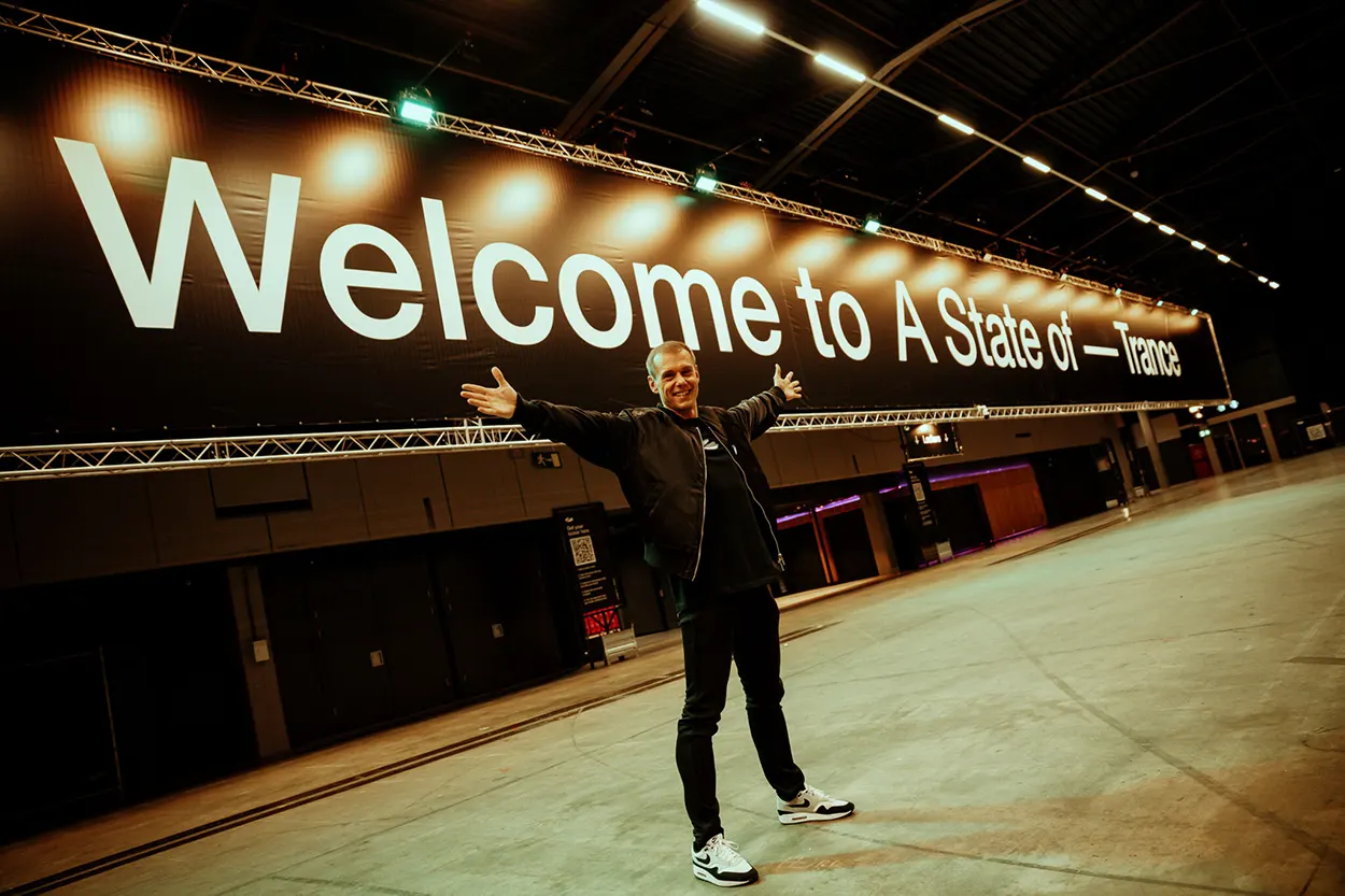 Armin van Buuren pose les bras ouverts devant une immense bannière « Welcome to A State Of Trance », dans une salle encore vide.