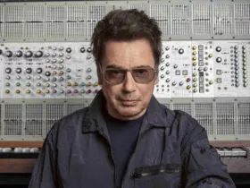 Jean-Michel Jarre pose devant un imposant synthétiseur modulaire, vêtu d’une veste sombre et portant des lunettes teintées.