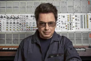 Jean-Michel Jarre pose devant un imposant synthétiseur modulaire, vêtu d’une veste sombre et portant des lunettes teintées.
