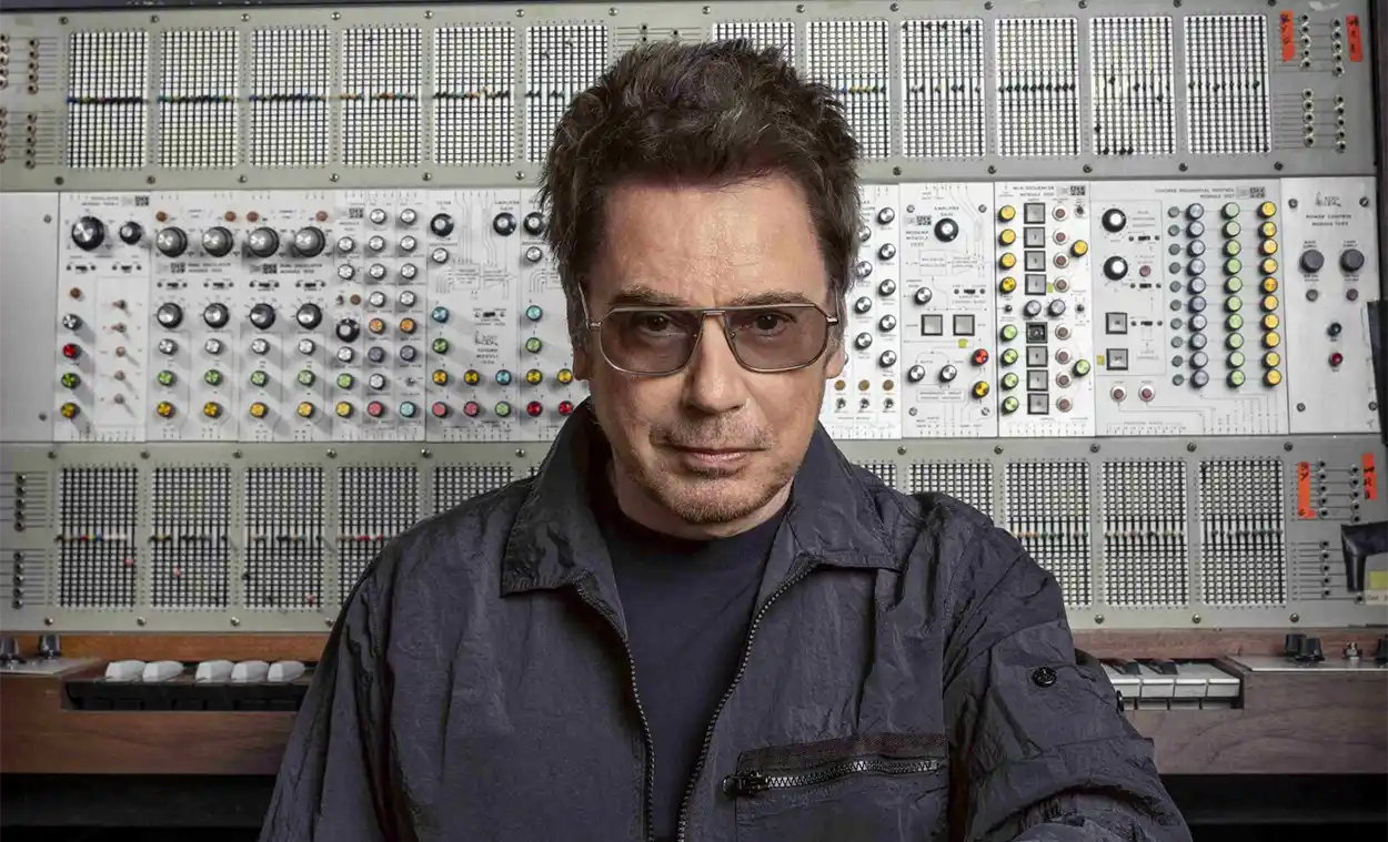 Jean-Michel Jarre pose devant un imposant synthétiseur modulaire, vêtu d’une veste sombre et portant des lunettes teintées.